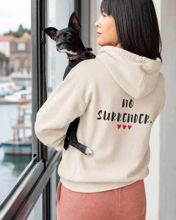 Back view of a woman wearing a beige hoodie featuring the inspirational quote “No Surrender” with red heart accents, holding a small dog by a window, motivational apparel promoting resilience, determination, strength, and perseverance.