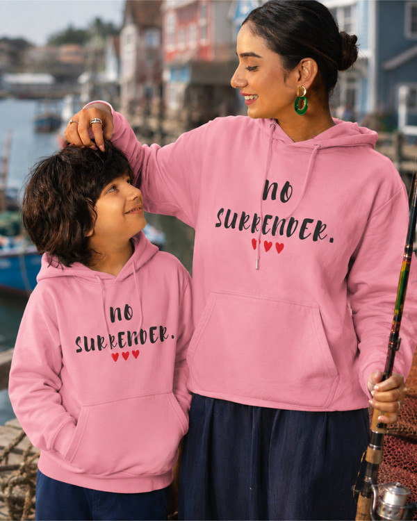 Woman and child wearing matching pink hoodies featuring the inspirational quote “No Surrender” with red heart accents, sharing a joyful moment outdoors, motivational apparel celebrating resilience, strength, perseverance, and hope.