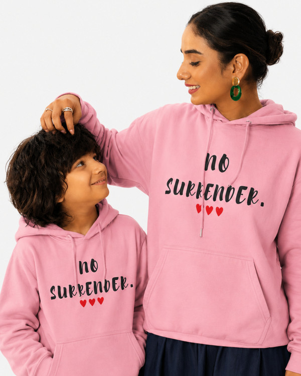 Woman and child wearing matching pink hoodies featuring the inspirational quote “No Surrender” with red heart accents, sharing a joyful moment outdoors, motivational apparel celebrating resilience, strength, perseverance, and hope.