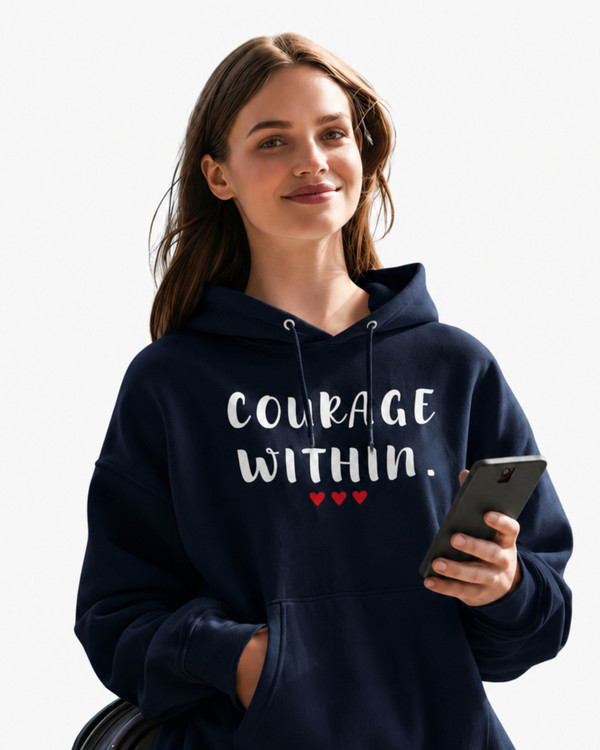 Woman wearing a navy blue Gildan 18500 hoodie featuring the “Courage Within” inspirational graphic, holding a smartphone and suitcase while standing in a bright airport terminal.