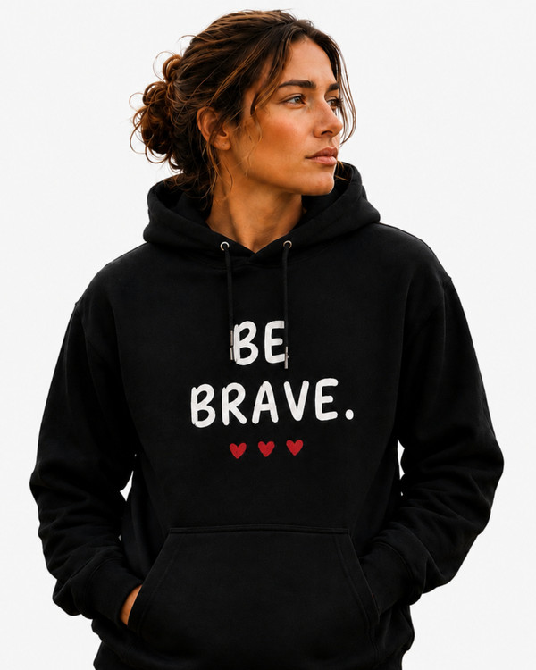 Woman modeling a navy blue motivational hoodie featuring the “BE BRAVE” graphic and three red hearts at a beach sunset. Inspirational apparel designed to encourage confidence, resilience, and self-belief.