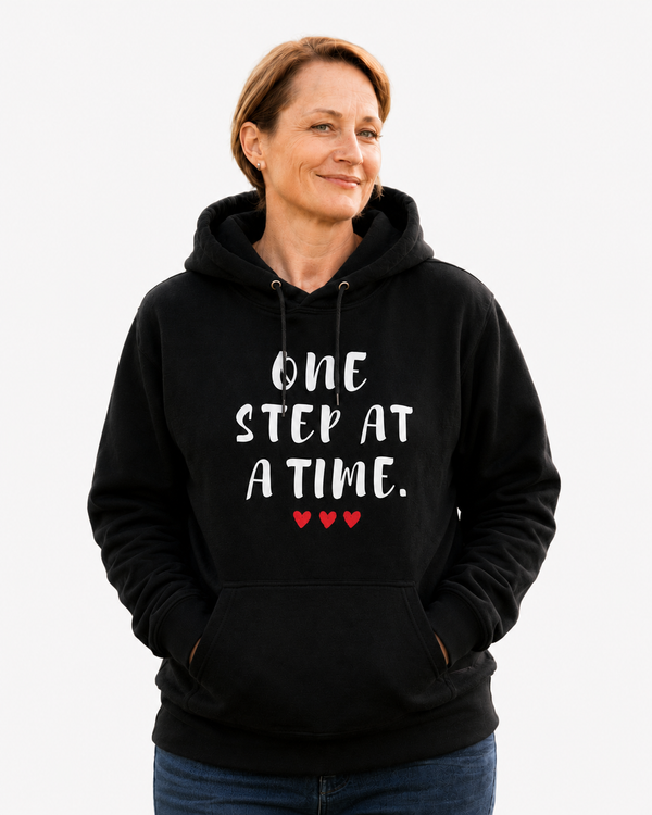 Woman wearing a black motivational hoodie with the quote “ONE STEP AT A TIME” and red hearts, helping someone up in a park, symbolizing encouragement, resilience, hope, and personal growth.