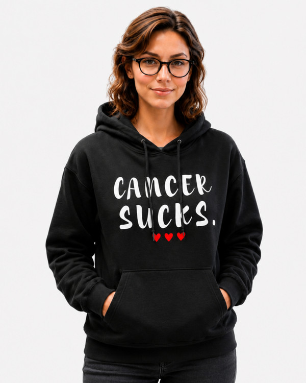 Woman wearing a black pullover hoodie featuring the phrase “Cancer Sucks” in distressed white lettering with three red hearts beneath. She is seated at a desk in a classroom or study environment, holding papers while looking toward the camera. The hoodie combines casual comfort with a powerful cancer awareness message of support, strength, and advocacy.