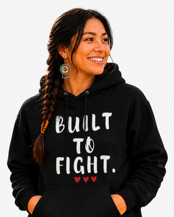Smiling woman wearing a black hoodie featuring the inspirational quote “Built to Fight” in distressed white lettering with red heart accents, standing on a tree-lined path during autumn.