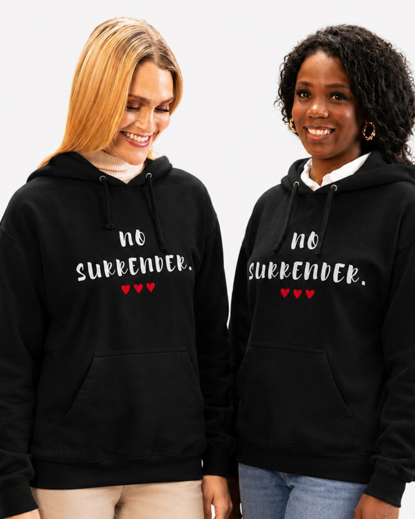 Two women wearing matching black hoodies featuring the inspirational quote “No Surrender” with red heart accents, smiling while baking together in a kitchen, motivational apparel celebrating resilience, determination, strength, and perseverance.