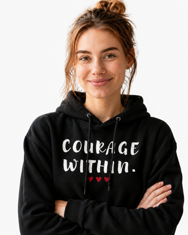 Woman wearing a black “Courage Within” motivational hoodie standing indoors with arms crossed in a bright, plant-filled home setting.