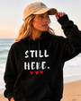 Female model wearing a black hoodie with the motivational phrase “STILL HERE.” and red heart accents. Meaningful sweatshirt promoting courage, determination, mental wellness, self-belief, and personal growth in a coastal setting.