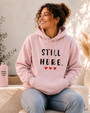Woman wearing a light pink hoodie featuring the empowering message “STILL HERE.” and three red hearts while seated indoors in a bright, cozy setting. Inspirational apparel symbolizing resilience, strength, perseverance, hope, and overcoming life's challenges.