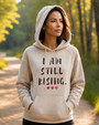Woman wearing a sand-colored inspirational hoodie featuring the message “I AM STILL RISING” with red heart accents, standing on a sunlit forest trail. Motivational women's apparel promoting resilience, personal growth, confidence, healing, and empowerment.