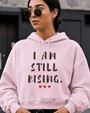Women's light pink motivational hoodie with “I AM STILL RISING” graphic and red heart details, photographed in a modern city environment. Inspirational apparel designed to celebrate strength, empowerment, recovery, determination, and positive mindset.