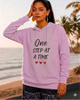 Woman standing near a beach at sunset wearing a light pink pullover hoodie featuring the inspirational phrase “One Step at a Time” in bold black lettering with three small red hearts beneath. The hoodie has a drawstring hood and relaxed fit, creating a comfortable and encouraging lifestyle look.