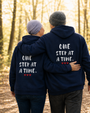 Two people walking arm in arm through a sunlit forest trail while wearing matching navy blue hoodies. The backs of the hoodies feature the inspirational phrase “One Step at a Time” in distressed white lettering with three small red hearts underneath. The image conveys support, companionship, resilience, and hope.