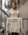 Woman wearing a sand-colored pullover hoodie featuring the supportive phrase “I Fight With You” in black distressed lettering with three red hearts beneath. A matching hoodie is displayed in the foreground. The hoodies feature a drawstring hood, front kangaroo pocket, and relaxed unisex fit. The design symbolizes encouragement, solidarity, strength, and standing together through life’s challenges.