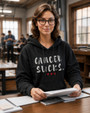 Woman wearing a black pullover hoodie featuring the phrase “Cancer Sucks” in distressed white lettering with three red hearts beneath. She is seated at a desk in a classroom or study environment, holding papers while looking toward the camera. The hoodie combines casual comfort with a powerful cancer awareness message of support, strength, and advocacy.