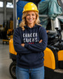 Woman wearing a navy blue pullover hoodie featuring the phrase “Cancer Sucks.” in distressed white lettering with three red hearts beneath. Standing confidently in an industrial workplace with a hard hat and forklift in the background, the image symbolizes strength, resilience, and support for cancer awareness. The hoodie combines comfort with a meaningful message of solidarity and hope.