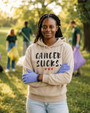 Young woman wearing a sand-colored pullover hoodie featuring the phrase “Cancer Sucks.” in distressed black lettering with three red hearts beneath. She stands outdoors in a park while participating in a community clean-up event. The hoodie combines a message of cancer awareness with themes of compassion, resilience, support, and positive action.