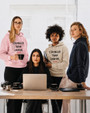 Four women gathered around a table in a bright, modern workspace. Three women are standing and one is seated behind a laptop. Two of the women are wearing hoodies featuring the inspirational message “STRONGER THAN CANCER.” in bold white lettering with small red hearts beneath the text. One hoodie is light pink and the other is sand beige. Coffee cups and camera equipment are arranged on the table, creating a collaborative and supportive atmosphere. The image conveys empowerment, unity, strength, and encouragement for those affected by cancer.