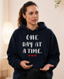 Woman seated indoors wearing a navy blue hoodie with the inspirational message “ONE DAY AT A TIME.” printed in large white lettering across the front, accented by three small red hearts beneath the text. She appears engaged in conversation or a support group discussion, gesturing with her hands while looking attentively to the side. The softly lit interior and calm expression create a warm, encouraging atmosphere. The design promotes hope, resilience, and taking life’s challenges one step at a time.