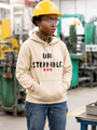 Person wearing a sand-colored pullover hoodie with the motivational message “UNSTOPPABLE.” printed in distressed black lettering and accented by three small red hearts beneath the text. They are standing in an industrial workshop while wearing a yellow hard hat and safety glasses, with their hands tucked into the hoodie’s kangaroo pocket. Machinery and manufacturing equipment are visible in the softly blurred background. The image conveys determination, resilience, confidence, and perseverance while showcasing the hoodie design in a hardworking, professional environment.