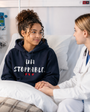 Young woman sitting in a hospital bed wearing a navy blue hoodie with the empowering message “UNSTOPPABLE.” printed in distressed white lettering and accented by three small red hearts beneath the text. She smiles warmly while holding hands with a healthcare professional seated beside her. The bright hospital room, medical equipment, and supportive interaction create a compassionate atmosphere focused on care, hope, and recovery. The image conveys resilience, strength, encouragement, and the determination to keep moving forward through life’s challenges.