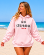Woman walking along a sunny beach wearing a light pink hoodie with the motivational message “UNSTOPPABLE.” printed in distressed black lettering and accented by three small red hearts beneath the text. She smiles while looking off into the distance, with the ocean, blue sky, and sandy shoreline creating a bright and uplifting coastal backdrop. The hoodie features a drawstring hood and kangaroo pocket, and the design is prominently displayed. The image conveys confidence, resilience, strength, and a positive outlook while showcasing the hoodie in a relaxed outdoor setting.