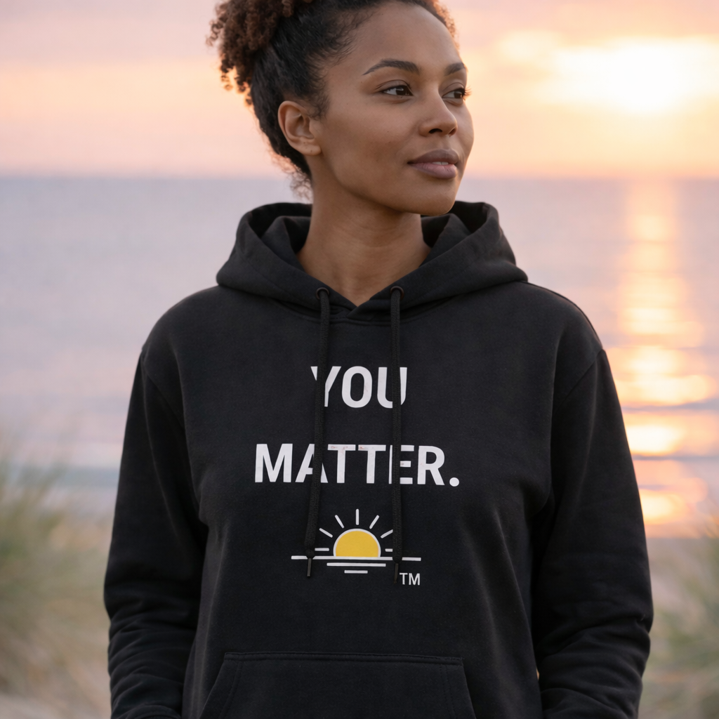 The image features a young woman wearing a black hoodie, smiling and walking outdoors. The background is softly blurred, with warm, golden-yellow tones suggestive of sunlight filtering through trees. This creates a sense of depth and focuses attention on the woman and the text overlaid on the image.  The main content of the image is the woman herself, who appears healthy and optimistic. She is positioned slightly off-center, with her gaze directed forward and to the left. The text, presented in white, is a prominent part of the composition, placed centrally and below the woman's chest.  The scene characteristics suggest an outdoor, natural setting, possibly a park or a wooded area, during daytime, perhaps late afternoon given the warm light. The focus on the woman and the motivational text indicates that this image is likely used for marketing or promotional purposes, possibly for athletic wear or a lifestyle brand.  The visual effects employed are a shallow depth of field, which blurs the background and isolates the subject, and the use of text overlay. The lighting is soft and appears to be natural, highlighting the woman's features and the texture of the hoodie. The overall mood is positive, empowering, and serene.  The text reads "Strength Collection" as a title, followed by a descriptive tagline: "A bold message of resilience. Wear it as a reminder that strength can rise even in life's toughest battles." This text reinforces the visual message of empowerment and inner strength conveyed by the woman's expression and the overall atmosphere of the image.