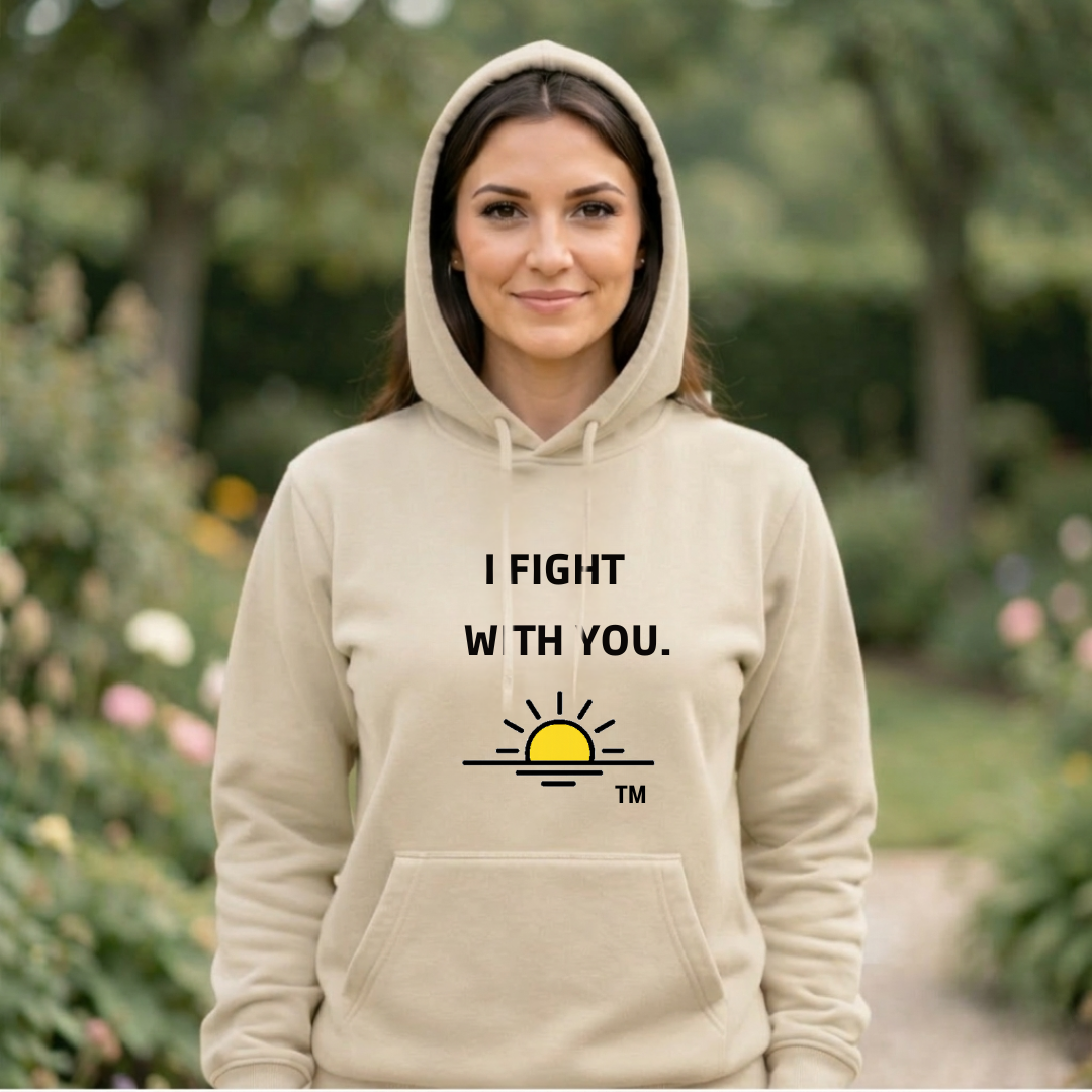 The image features a woman from the chest up, wearing a dark navy hooded sweatshirt. She has medium-length brown hair and is looking directly at the camera with a gentle smile. The background is blurred, suggesting an outdoor setting with trees and foliage, bathed in warm, diffused light, possibly from late afternoon sun. The overall mood is calm and inviting.  Overlaid on the image, towards the lower half, are two lines of white text. The first line reads "Support Collection" in a larger, bolder font. Beneath it, in a smaller font, is the text "A powerful message of solidarity for those standing beside someone they love.". This text suggests the image is part of a promotional campaign or a message related to a collection of items designed to convey support and solidarity.  The visual effects primarily involve the use of shallow depth of field to isolate the subject and create a soft, atmospheric background. The lighting is soft and warm, highlighting the subject's face and hair. The composition places the woman slightly off-center, with her gaze directly engaging the viewer. The text is strategically placed to not obscure the main subject but to complement the overall message. The contrast between the dark sweatshirt and the lighter background helps the subject stand out.