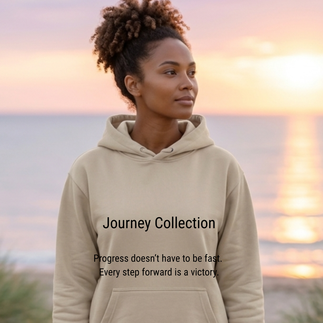 The image displays a medium shot portrait of a young Black woman with curly hair, wearing a light beige hooded sweatshirt. She is looking off to the right side of the frame with a thoughtful expression. The background consists of a serene beach scene at sunset or sunrise. The sky is a soft gradient of pinks, oranges, and purples, with the calm ocean stretching out to the horizon. A shimmering reflection of the sky's colors is visible on the water. The overall scene is tranquil and peaceful, evoked by the soft lighting and the natural setting.  Text overlays are present on the sweatshirt. The upper text reads "Journey Collection" in a prominent, dark font. Below this, in a slightly smaller font, is the motivational quote "Progress doesn't have to be fast. Every step forward is a victory."  The visual effects contribute to a warm and inspiring mood. The soft focus on the background draws attention to the woman and the text. The colors of the sunset are warm and inviting, enhancing the overall aesthetic. The placement of the text is centered and clear, making it easily readable against the lighter fabric of the sweatshirt. The composition is balanced, with the woman positioned slightly off-center, creating visual interest. The lighting is gentle, suggesting either dawn or dusk, which often symbolizes new beginnings or moments of reflection.