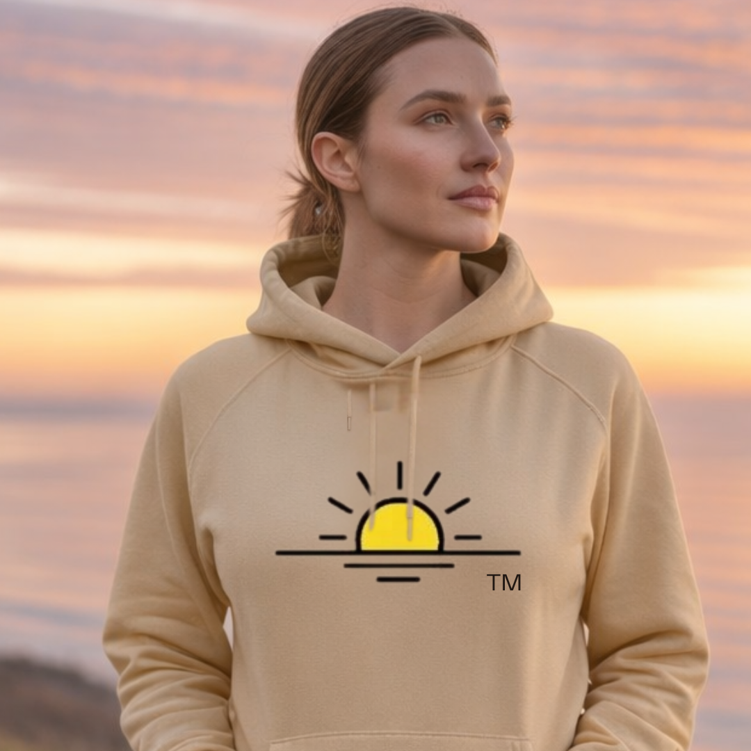 The image features a woman from the chest up, wearing a dark navy hooded sweatshirt. She has medium-length brown hair and is looking directly at the camera with a gentle smile. The background is blurred, suggesting an outdoor setting with trees and foliage, bathed in warm, diffused light, possibly from late afternoon sun. The overall mood is calm and inviting.  Overlaid on the image, towards the lower half, are two lines of white text. The first line reads "Support Collection" in a larger, bolder font. Beneath it, in a smaller font, is the text "A powerful message of solidarity for those standing beside someone they love.". This text suggests the image is part of a promotional campaign or a message related to a collection of items designed to convey support and solidarity.  The visual effects primarily involve the use of shallow depth of field to isolate the subject and create a soft, atmospheric background. The lighting is soft and warm, highlighting the subject's face and hair. The composition places the woman slightly off-center, with her gaze directly engaging the viewer. The text is strategically placed to not obscure the main subject but to complement the overall message. The contrast between the dark sweatshirt and the lighter background helps the subject stand out.