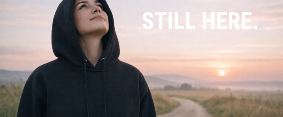 Woman in black hoodie facing sunset overlooking river STILL HERE. Strength. Courage Hope