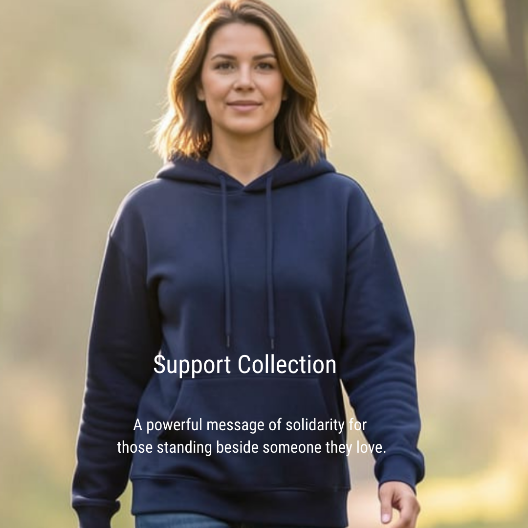 The image features a woman from the chest up, wearing a dark navy hooded sweatshirt. She has medium-length brown hair and is looking directly at the camera with a gentle smile. The background is blurred, suggesting an outdoor setting with trees and foliage, bathed in warm, diffused light, possibly from late afternoon sun. The overall mood is calm and inviting.  Overlaid on the image, towards the lower half, are two lines of white text. The first line reads "Support Collection" in a larger, bolder font. Beneath it, in a smaller font, is the text "A powerful message of solidarity for those standing beside someone they love.". This text suggests the image is part of a promotional campaign or a message related to a collection of items designed to convey support and solidarity.  The visual effects primarily involve the use of shallow depth of field to isolate the subject and create a soft, atmospheric background. The lighting is soft and warm, highlighting the subject's face and hair. The composition places the woman slightly off-center, with her gaze directly engaging the viewer. The text is strategically placed to not obscure the main subject but to complement the overall message. The contrast between the dark sweatshirt and the lighter background helps the subject stand out.