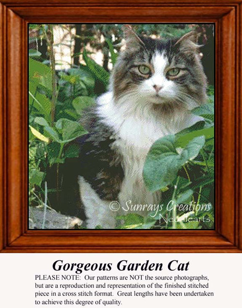 A black, white, and gray cat with green eyes sits among lush green plants in a garden, ready for your next cross stitch project.