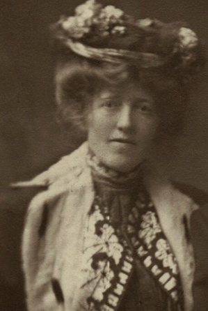A 19th-century photograph showing a bust portrait of the painter Laura Theresa Alma-Tadema. She has dark, curly hair and is wearing an elegant Victorian-style dress with a high collar and decorative lace. Her expression is calm and creative, capturing the spirit of a leading female artist in the Pre-Raphaelite circle.