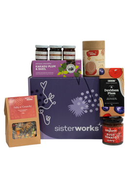 An overhead view of the open SisterWorks Foodie Hamper. The contents are neatly arranged in a box, including a jar of Eggplant Pickle, a 3-pack of ShortHive Infused Honey, a pouch of Cinnamon Myrtle Black Tea, a bag of Salty n' Crunchy Mix, and a box of Kakadu Plum Co. Crackers. The collection highlights a perfect gourmet gift set for a foodie