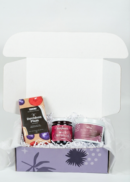An overhead view of the open SisterWorks Berry Sweet Hamper. The box contains a jar of SisterWorks Raspberry Jam, a SisterWorks Cherry Blossom Soy Candle, and a bar of BSKT Davidson Plum Vegan Chocolate. The items are arranged to showcase a sweet and fragrant gift set