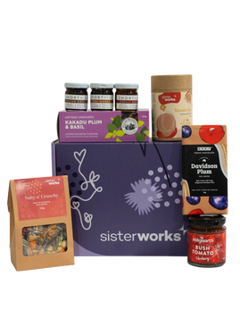 A SisterWorks Aussie Holiday Hamper. The box contains STREAT Gingerbread Stars, SisterWorks Raspberry Jam, BSKT Davidson Plum Vegan Chocolate, SisterWorks Lemon Myrtle Green Tea, and Australian Icon Coasters. The items are arranged neatly in a box, highlighting a festive and ethical gift set.