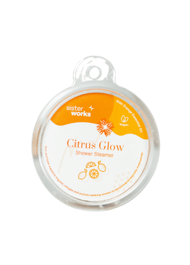 Front view of the SisterWorks Citrus Glow Shower Steamer in clear round packaging against a plain background. The orange and white label highlights 'Citrus Glow Shower Steamer,' 'With Orange Essential Oil,' and 'Vegan,' with text indicating support for migrant, refugee, and asylum-seeking women Front view of the SisterWorks Citrus Glow Shower Steamer in clear round packaging against a plain background. The orange and white label highlights 'Citrus Glow Shower Steamer,' 'With Orange Essential Oil,' and 'Vegan,' with text indicating support for migrant, refugee, and asylum-seeking women