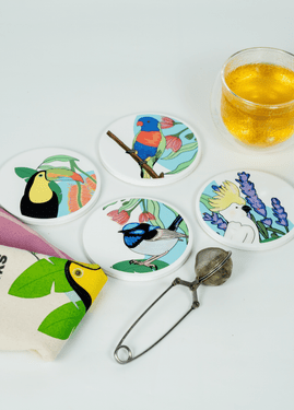 Overhead view of four round SisterWorks Cultural Fusion Coasters, each featuring a unique vibrant illustration of birds and plants by artist Marynel Jaimes. A SisterWorks tea towel with a toucan design and a glass of tea with an infuser are also visible on a white background, highlighting a cultural and artistic gift set.