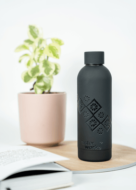 Side view of the black 'Embroidered Roots' reusable bottle by SisterWorks on a wooden coaster, next to an open book and a potted plant in a light-filled setting. The intricate 'Embroidered Roots' design by Fernanda Rodriguez is prominent on the bottle, showcasing a stylish and sustainable everyday item. Side view of the black 'Embroidered Roots' reusable bottle by SisterWorks on a wooden coaster, next to an open book and a potted plant in a light-filled setting. The intricate 'Embroidered Roots' design by Fernanda Rodriguez is prominent on the bottle, showcasing a stylish and sustainable everyday item.