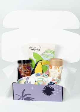 Angled view of the SisterWorks Cultural Fusion Hamper contents neatly arranged within the purple standard gift box. Features SisterWorks Eggplant Pickle, Masala Chai, a toucan-designed Cultural Fusion Tea Towel, and four vibrant Cultural Fusion Coasters by artist Marynel Jaimes, representing a thoughtful and ethically sourced cultural gift. Angled view of the SisterWorks Cultural Fusion Hamper contents neatly arranged within the purple standard gift box. Features SisterWorks Eggplant Pickle, Masala Chai, a toucan-designed Cultural Fusion Tea Towel, and four vibrant Cultural Fusion Coasters by artist Marynel Jaimes, representing a thoughtful and ethically sourced cultural gift.