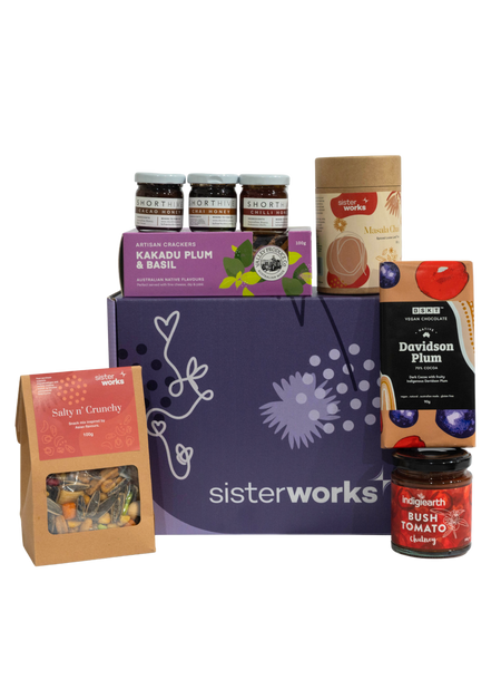 An overhead view of the open SisterWorks Foodie Hamper. The contents are neatly arranged in a box, including a jar of Eggplant Pickle, a 3-pack of ShortHive Infused Honey, a pouch of Cinnamon Myrtle Black Tea, a bag of Salty n' Crunchy Mix, and a box of Kakadu Plum Co. Crackers. The collection highlights a perfect gourmet gift set for a foodie