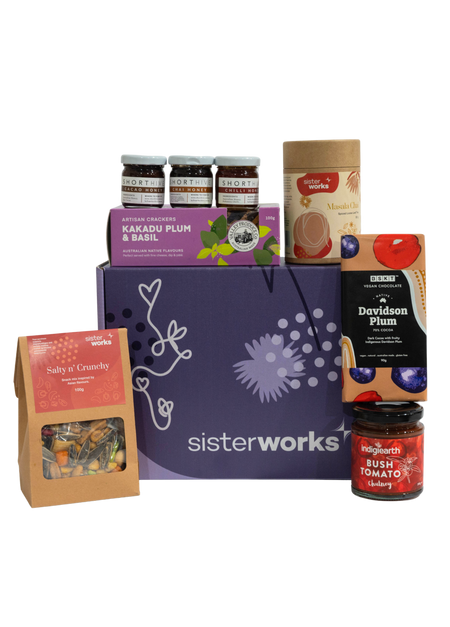 A SisterWorks Aussie Holiday Hamper. The box contains STREAT Gingerbread Stars, SisterWorks Raspberry Jam, BSKT Davidson Plum Vegan Chocolate, SisterWorks Lemon Myrtle Green Tea, and Australian Icon Coasters. The items are arranged neatly in a box, highlighting a festive and ethical gift set.