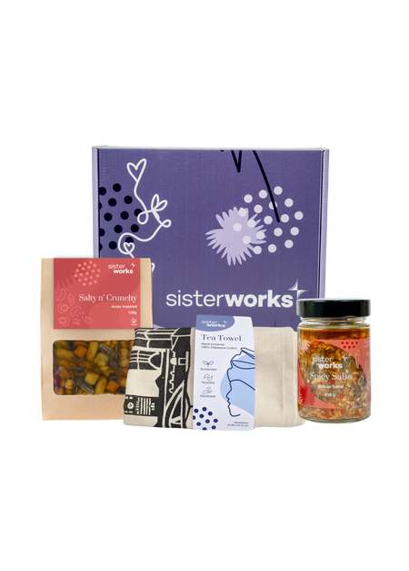 An overhead view of the open SisterWorks Savoury Treats Hamper. The box contains a colourful tea towel, a jar of SisterWorks Spicy Salsa, and a bag of Salty n' Crunchy Mix, all arranged to showcase a collection of gourmet, savoury snacks.