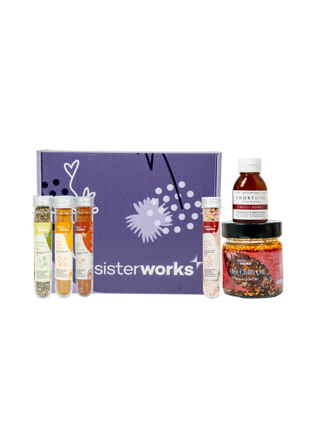 An overhead view of the open SisterWorks Spice Enthusiast's Delight Hamper. The hamper contains a collection of gourmet spices, including Hot Chilli Oil, Smoky Herb Seasoning, Zesty Spice Blend, Herbal Lemon Mix, and Spicy Fusion Salt. The items are arranged in a box, highlighting a beautiful gift set for anyone who loves to cook.