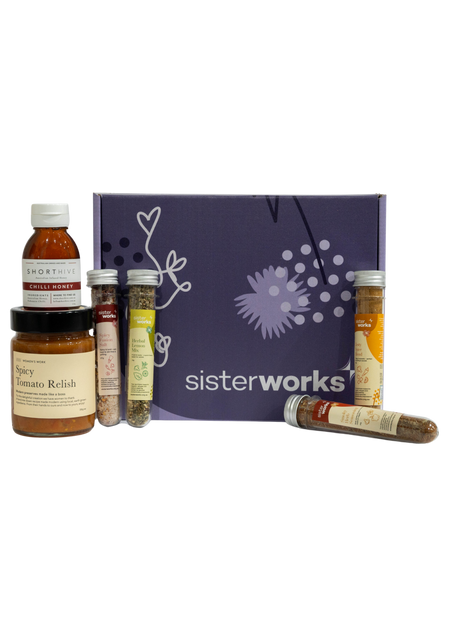 An overhead view of the open SisterWorks Spice Enthusiast's Delight Hamper. The hamper contains a collection of gourmet spices, including Hot Chilli Oil, Smoky Herb Seasoning, Zesty Spice Blend, Herbal Lemon Mix, and Spicy Fusion Salt. The items are arranged in a box, highlighting a beautiful gift set for anyone who loves to cook.