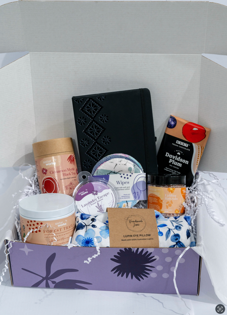 An overhead view of the open SisterWorks Wellbeing Hamper. The contents are neatly arranged inside a box, including a SisterWorks Premium Hard Cover Journal, a pen, an eye pillow, a bar of BSKT Chocolate, a pouch of Cinnamon Myrtle Black Tea, a container of Uplifting Bath Salts, a Shower Steamer Lavender Escape, and an Australian Flowers Candle, highlighting a collection of self-care and journaling products.