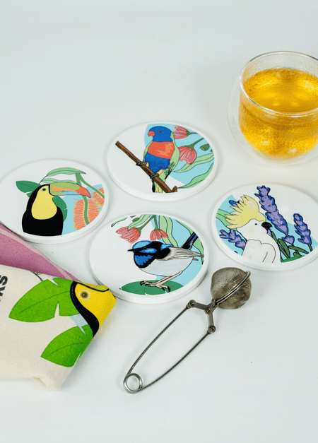 Overhead view of four round SisterWorks Cultural Fusion Coasters, each featuring a unique vibrant illustration of birds and plants by artist Marynel Jaimes. A SisterWorks tea towel with a toucan design and a glass of tea with an infuser are also visible on a white background, highlighting a cultural and artistic gift set.