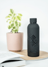 Side view of the black 'Embroidered Roots' reusable bottle by SisterWorks on a wooden coaster, next to an open book and a potted plant in a light-filled setting. The intricate 'Embroidered Roots' design by Fernanda Rodriguez is prominent on the bottle, showcasing a stylish and sustainable everyday item.