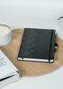 Overhead view of the black 'Embroidered Roots' Hard Cover Journal by SisterWorks, resting on a wooden surface next to a lit candle and an open book. The intricate 'Embroidered Roots' pattern, designed by Fernanda Rodriguez, is visible on the journal's cover, showcasing a sophisticated and inspiring tool for daily writing