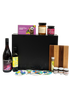 Front view of the SisterWorks Artisan Grazing Hamper contents laid out. Features Indigiearth Lemon Myrtle Olive Oil, Munificent Wine, ShortHive Infused Honey, SisterWorks Spicy Salsa, SisterWorks Fig Jam, Kakadu Plum & Basil Crackers, Cultural Fusion Coasters, and a Nigretta of Hamilton wooden board. The black premium gift box with embossed SisterWorks logo is visible in the background, signifying a luxury, ethical corporate gift